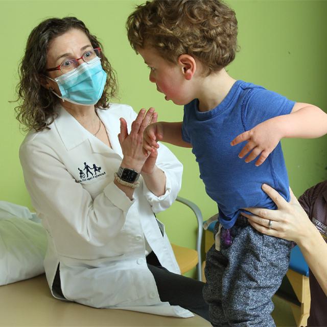 一个穿着蓝色衬衫、一头卷发的蹒跚学步的孩子被一位理疗师抱着，他举起双臂, 史黛西·萨斯考尔在一旁观看. 